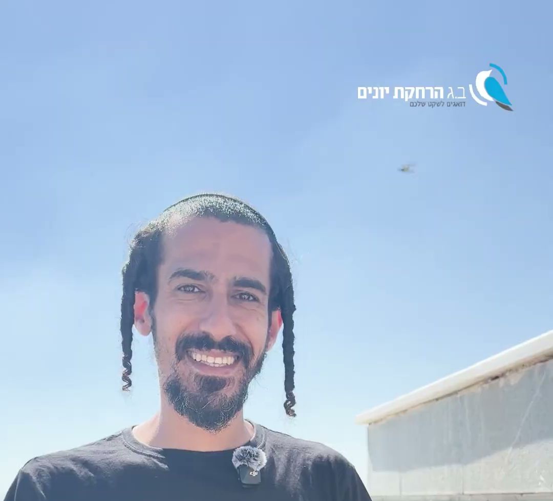 טכנאי מחייך עומד ליד מעקה גג, ציפור בתעופה בשמיים מאחוריו