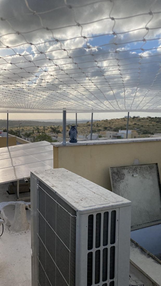 רשת מתוחה על עמודים מעל גג טכני עם מעבה מיזוג ופאנל, ועל המעקה פסל ינשוף פלסטיק